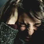 Close-up of a woman in distress with eyes closed and hands in hair, expressing anxiety.