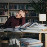 A tired woman surrounded by books, studying in a library, feeling overworked and stressed.