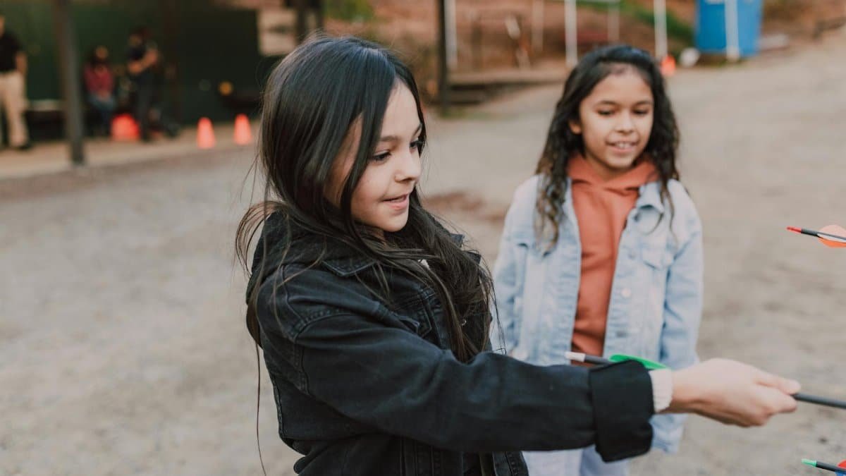 Two young girls engaging in outdoor archery practice, having fun under supervision.