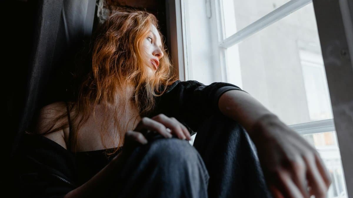 A woman with red hair sits by a window, deep in thought, conveying emotion and reflection.