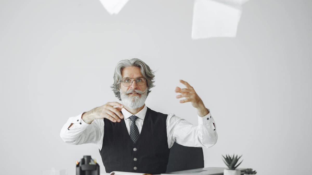 Elderly man in formal attire throwing papers at modern desk, symbolizing stress or creativity.
