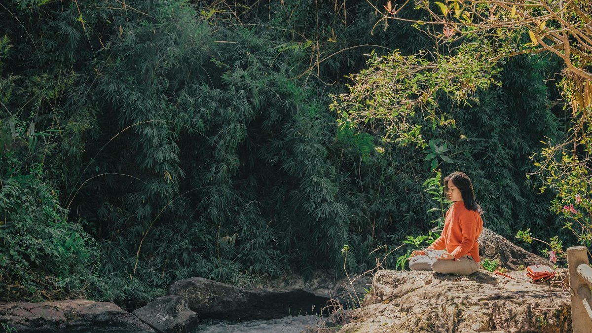 Woman meditating by a serene riverside surrounded by lush greenery on a sunny day.