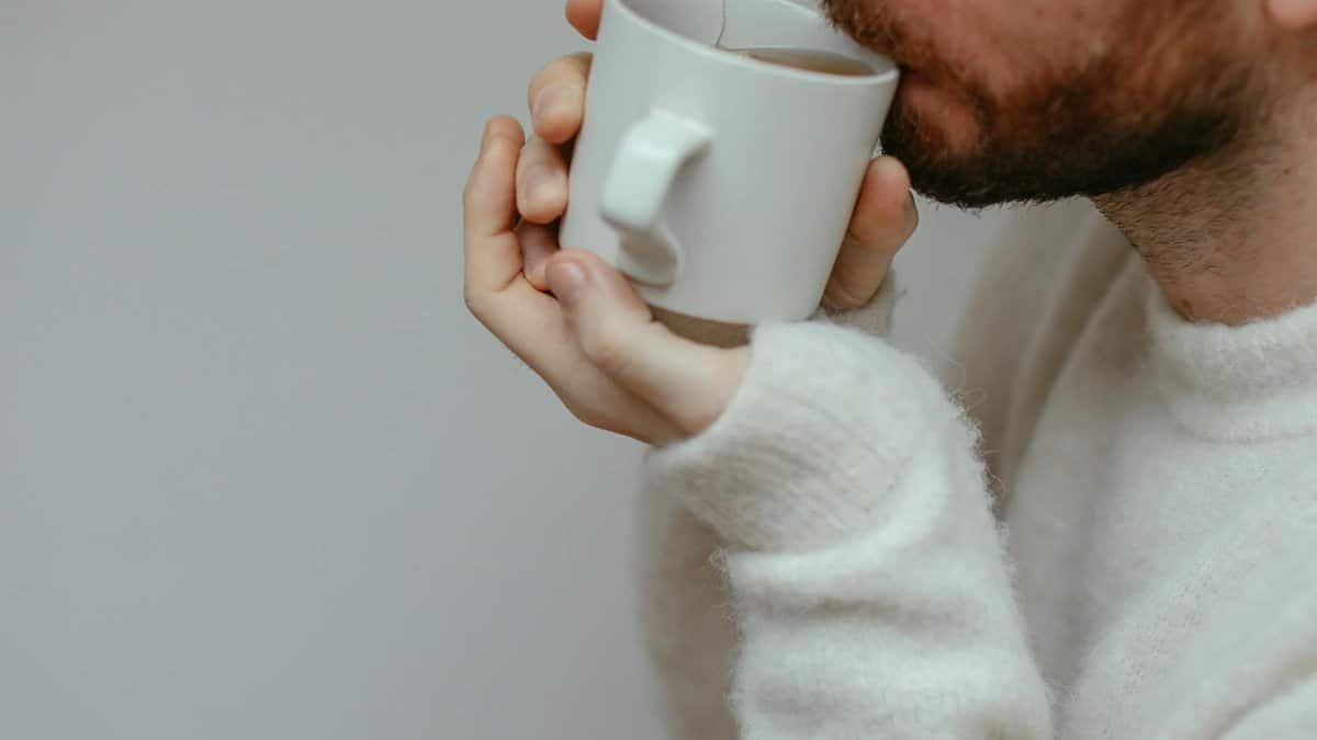 Close-up of a bearded man sipping a hot drink, wearing a cozy sweater indoors. Warm and inviting setting.