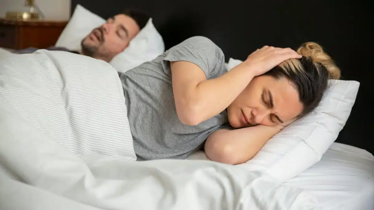 A woman looks frustrated in bed as her partner snores loudly, highlighting sleep disturbance issues.