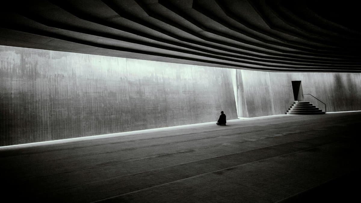 A lone person meditates in a vast, modern, minimalist indoor hall with dramatic architecture.