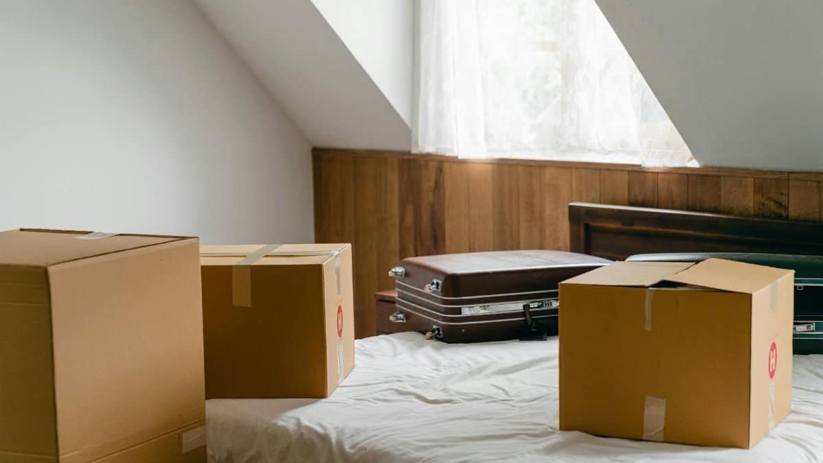 Small cardboard boxes and leather cases placed on bed with sheet near big box in light room of house under sloping roof