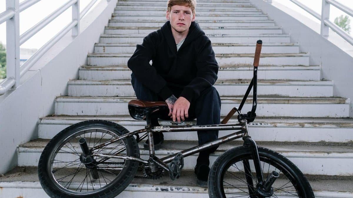 A young man in a black sweater sitting on stairs with a BMX bike in an urban setting.