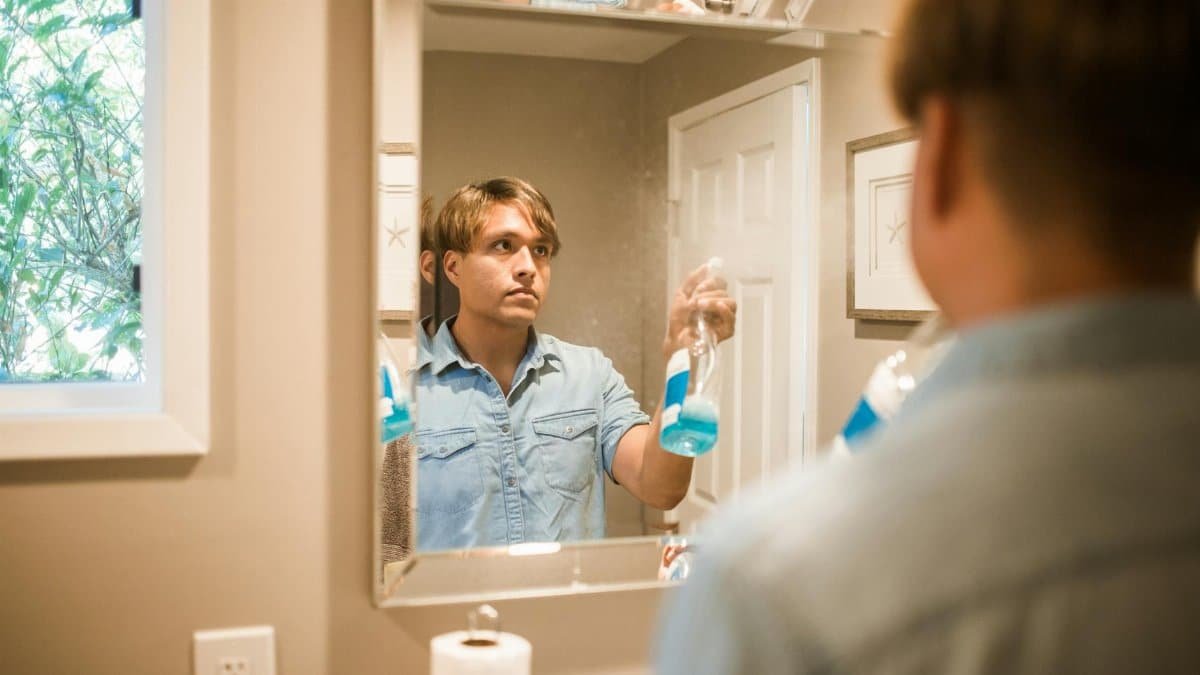 Man cleaning bathroom mirror with blue spray bottle reflection in the frame.
