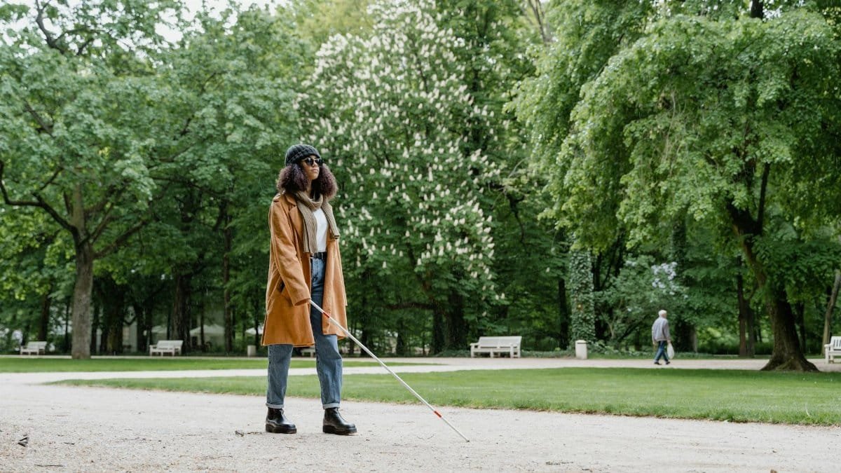A blind woman using a white cane walks through a lush green park on a sunny day.