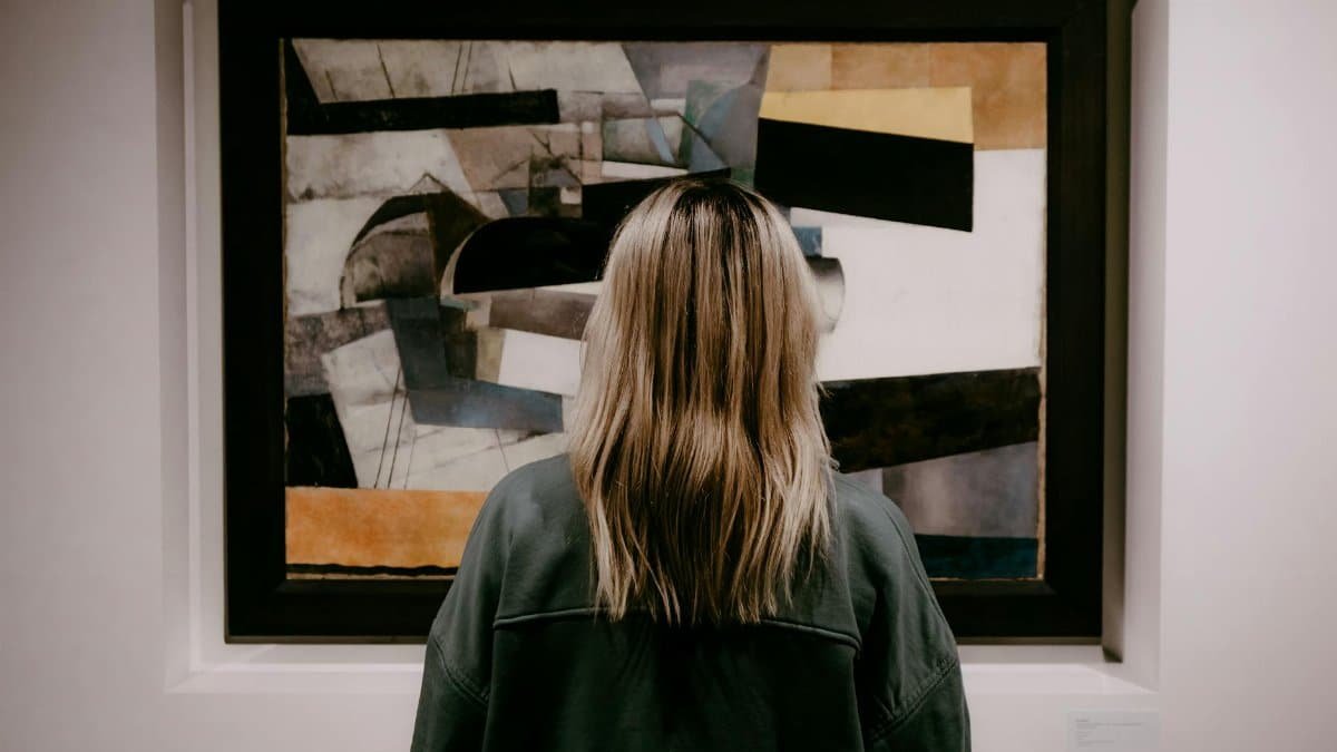 Back view of a woman with blonde hair viewing an abstract painting in an art gallery.