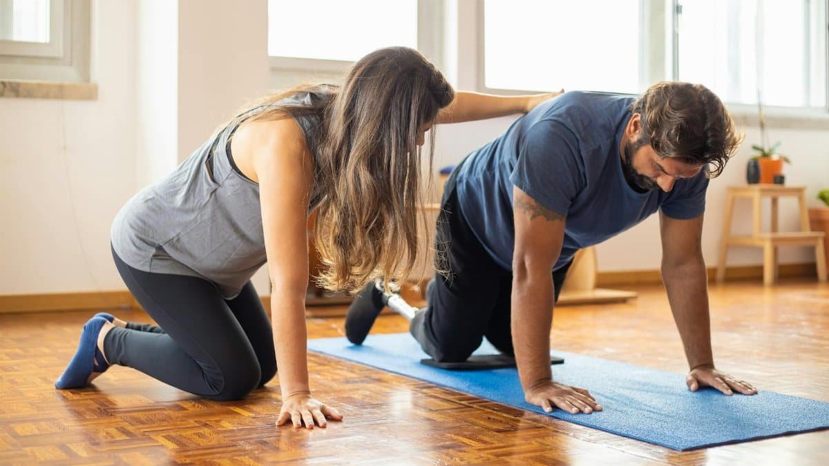 A man with a prosthetic leg exercises with a trainer indoors for adaptive fitness.