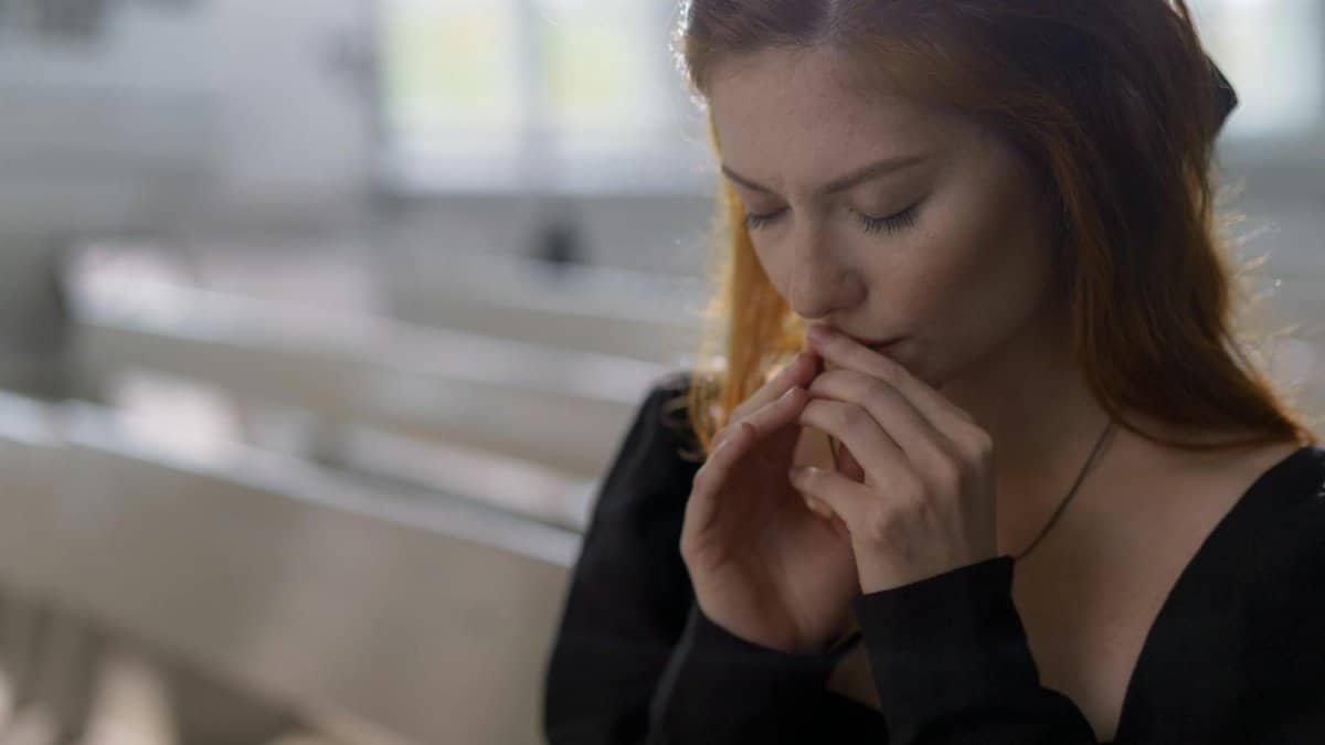 A young woman with eyes closed prays thoughtfully in a sunlit church.