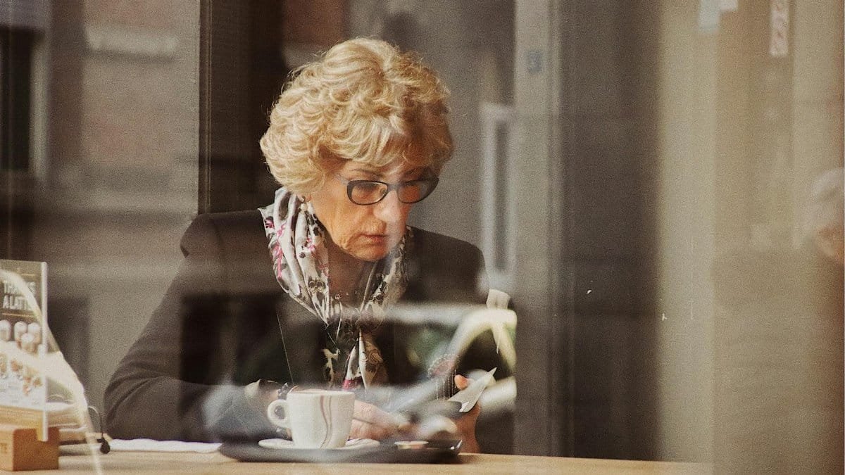 Senior woman in a cafe, reading with coffee, captured through a window reflection, emphasizing solitude and concentration.