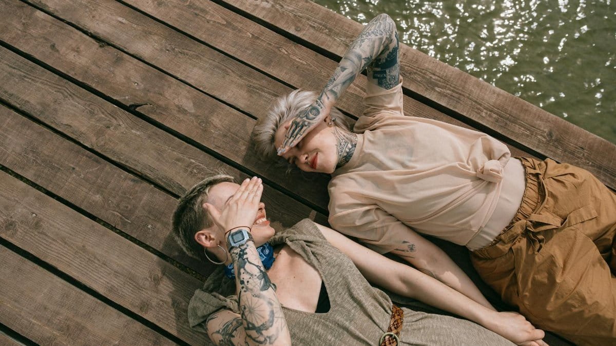 Two women with tattoos relax on a wooden deck by the water, embodying friendship.