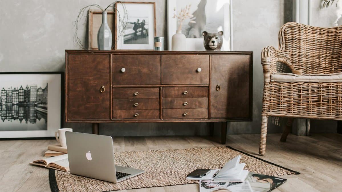 A cozy home office interior featuring a rustic wooden cabinet, wicker chair, and open laptop.