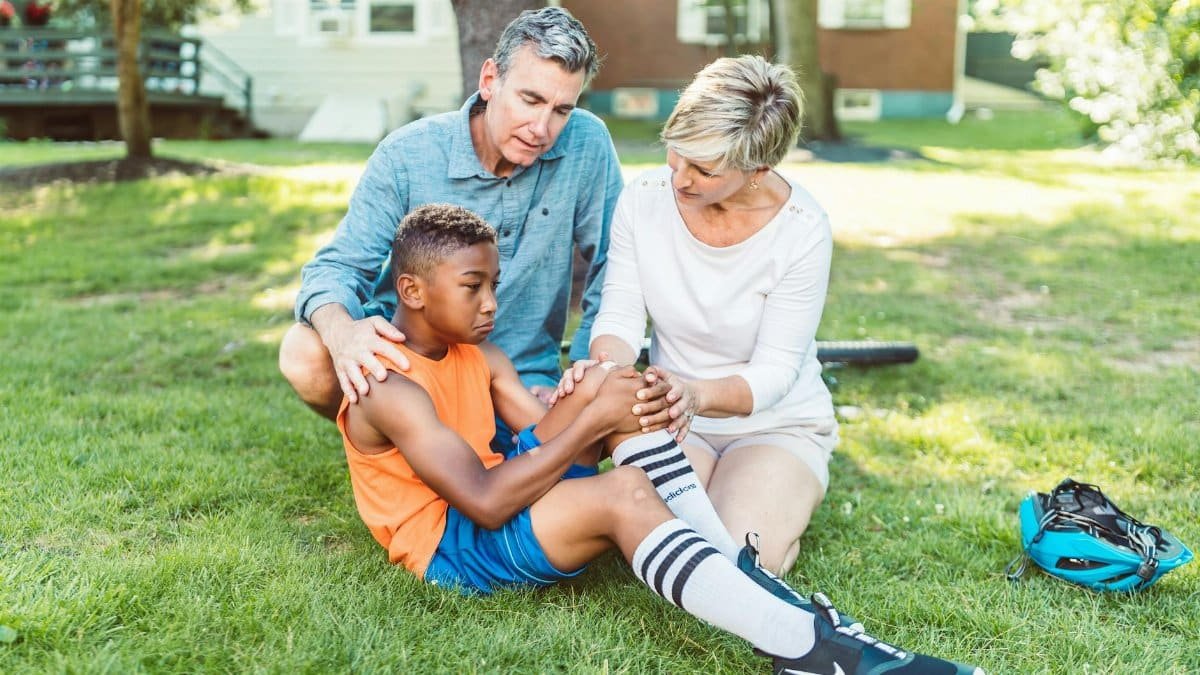 Parents comforting their injured son sitting on the grass in a backyard setting. Daytime scene.