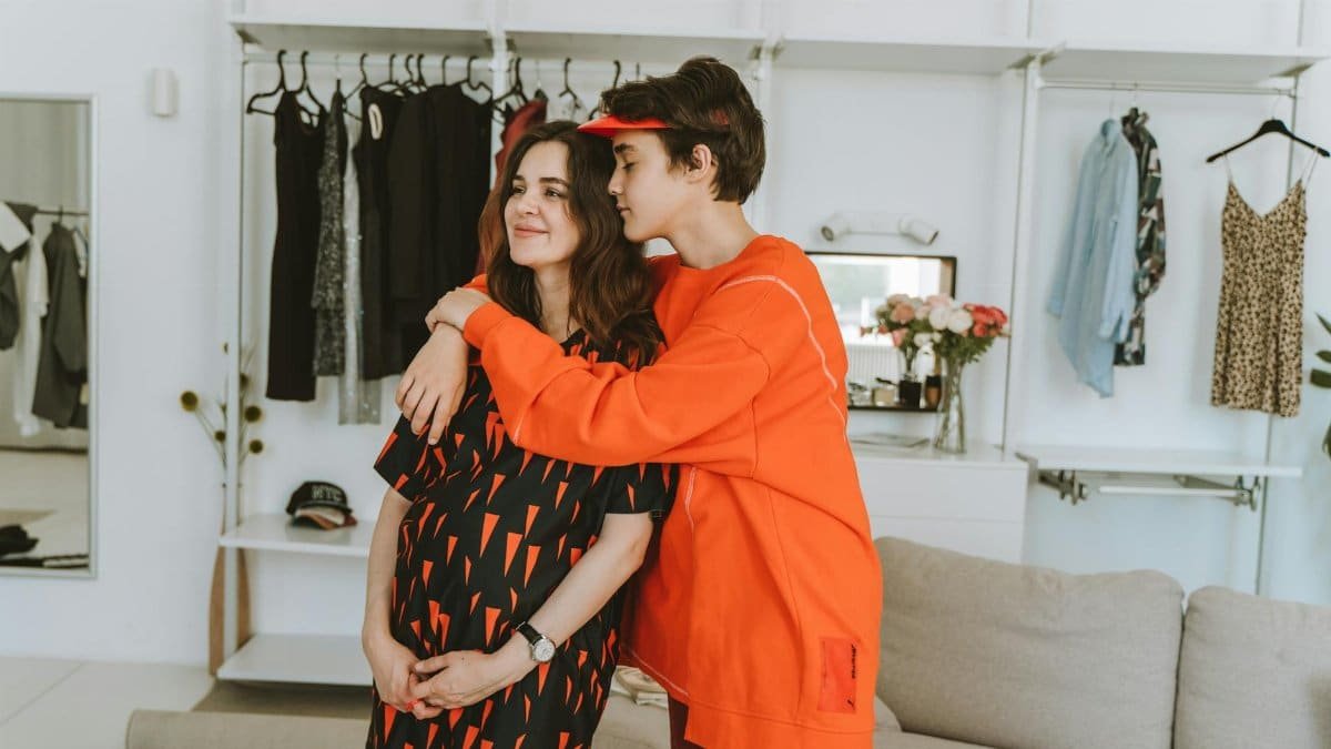 Pregnant woman and teenage son share an affectionate hug in a stylish indoor clothing store setting.