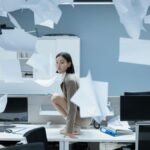 Woman perched on a desk in a chaotic office with papers flying, representing workplace stress.