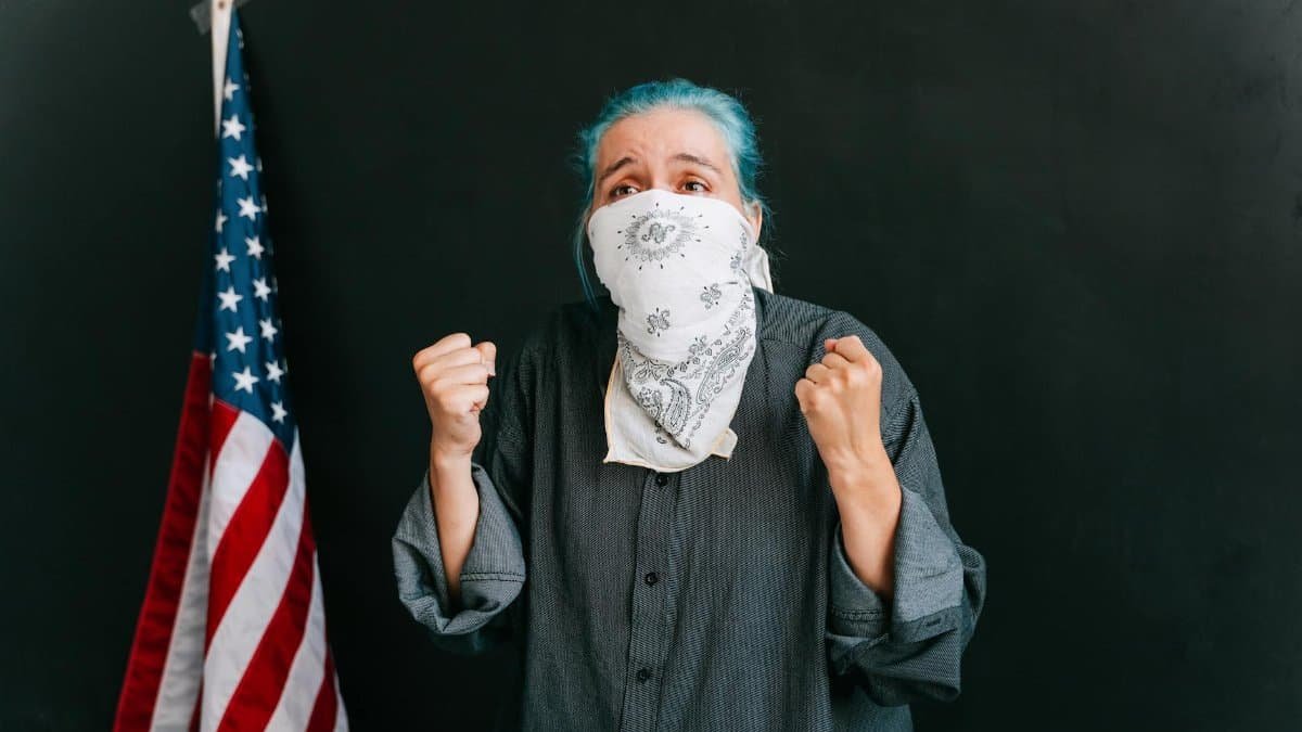 A woman passionately protesting with clenched fists, wearing a bandana mask, next to an American flag.