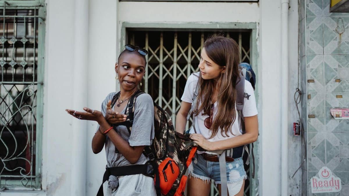 Two women stand laughing with backpacks, exploring a city during their travel adventure.