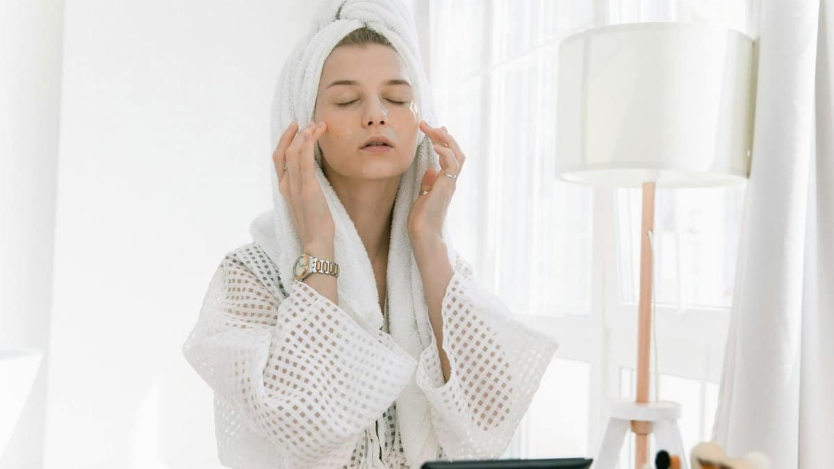Adult woman in bathrobe doing skincare routine with head towel and under-eye patches indoors.