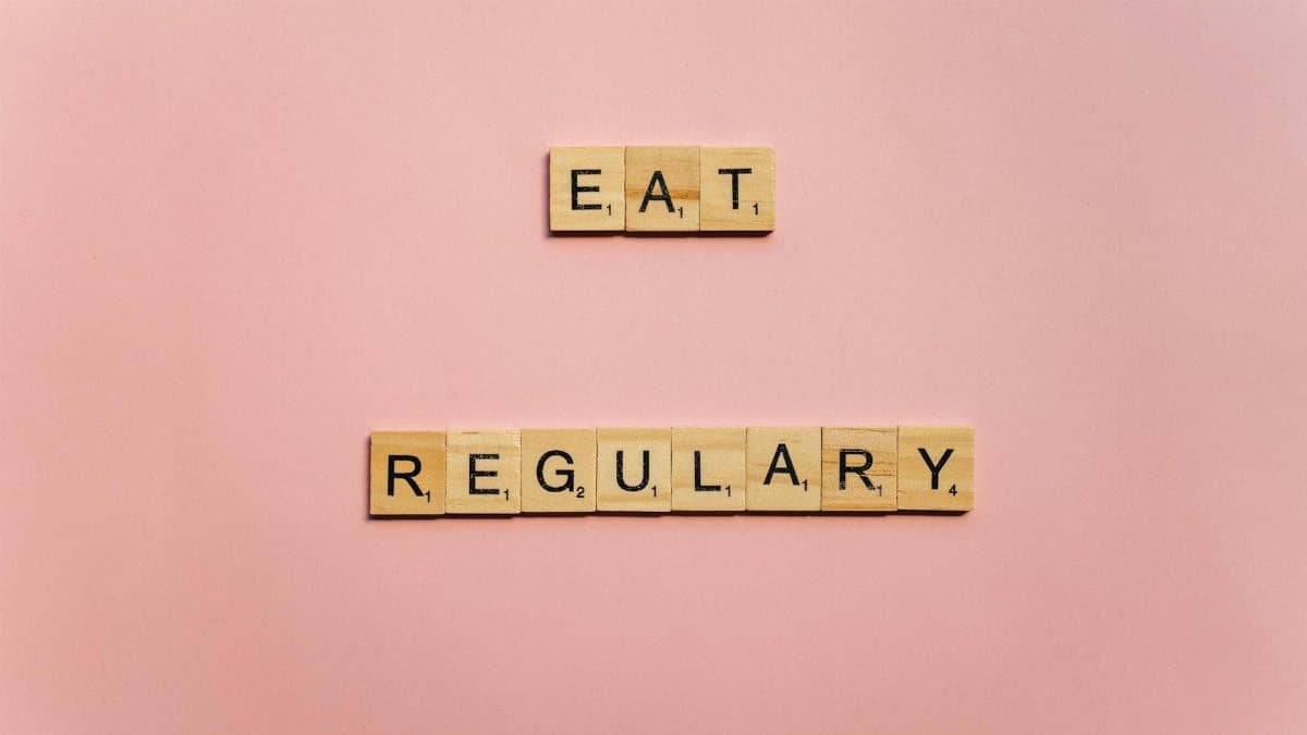 Wooden tiles spelling 'Eat Regularly' on a pink background, promoting healthy eating habits.