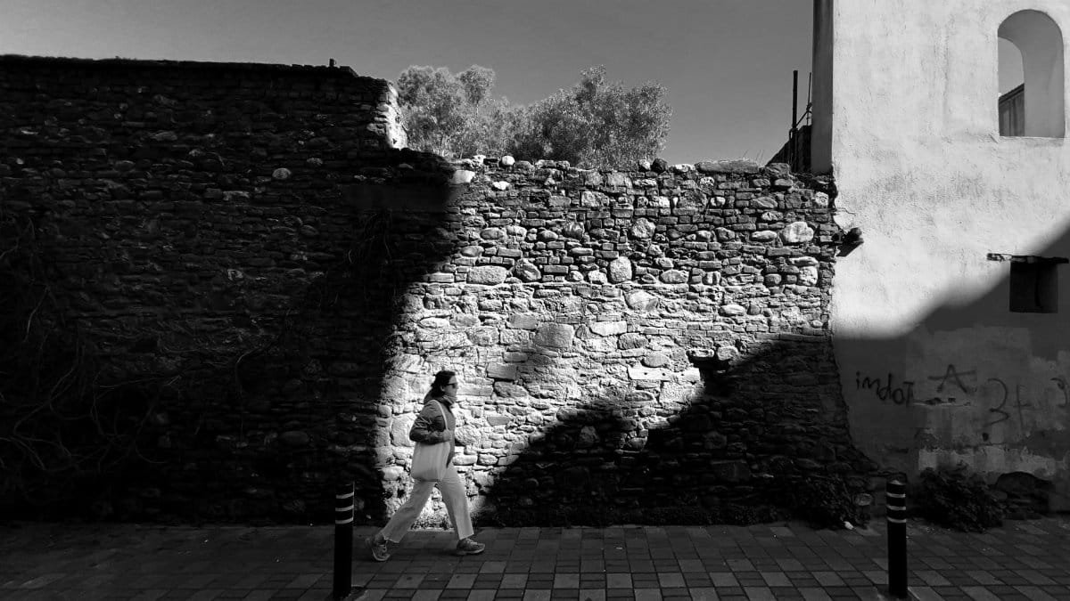 Black and white photo of a woman walking past a historic stone wall in Aydın, Türkiye.