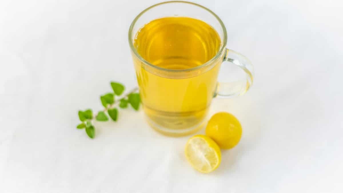 A clear glass of lemon herbal tea accompanied by fresh herbs on a white surface, offering a refreshing drink.