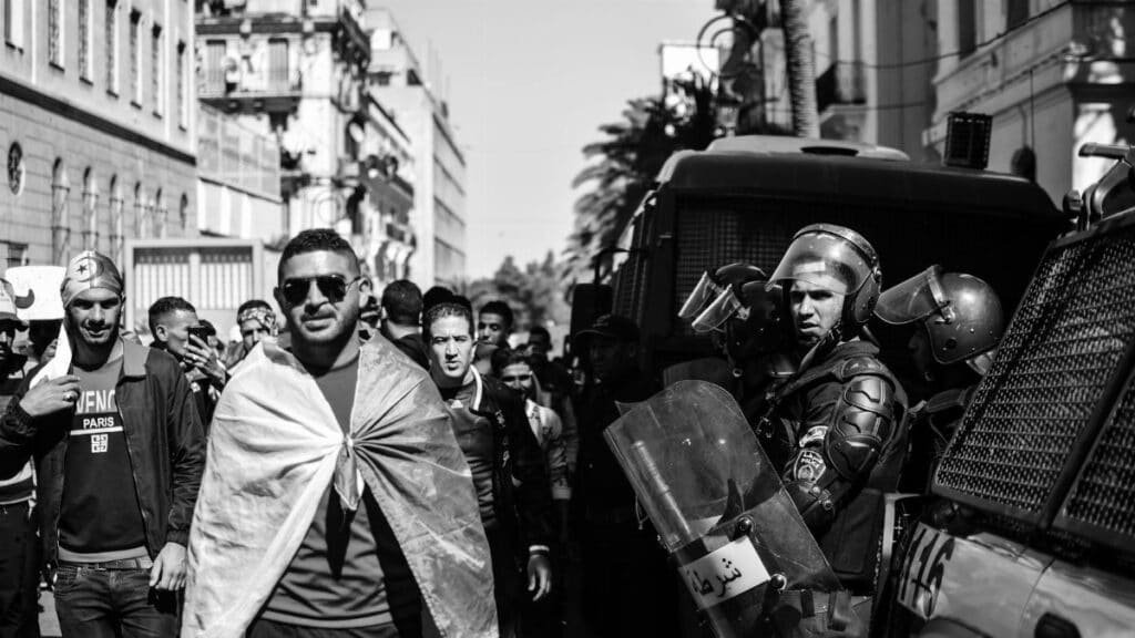 Black and white scene of protesters and police in an Algerian street, highlighting tension.