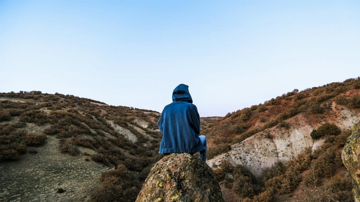 A solitary figure in a hoodie sits on a rocky hill overlooking a landscape at dusk, embracing nature's serenity.
