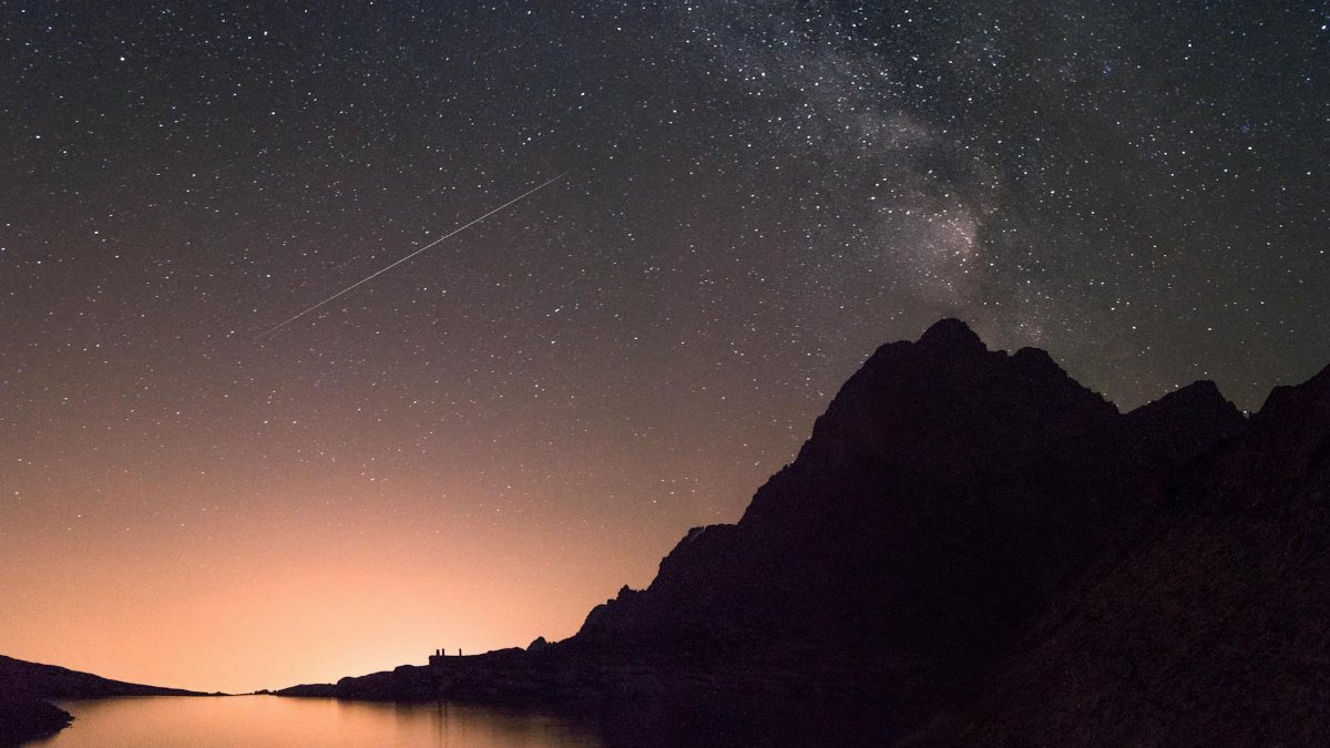 A captivating view of the Milky Way and shooting star above a silhouetted mountain reflecting over a calm lake.