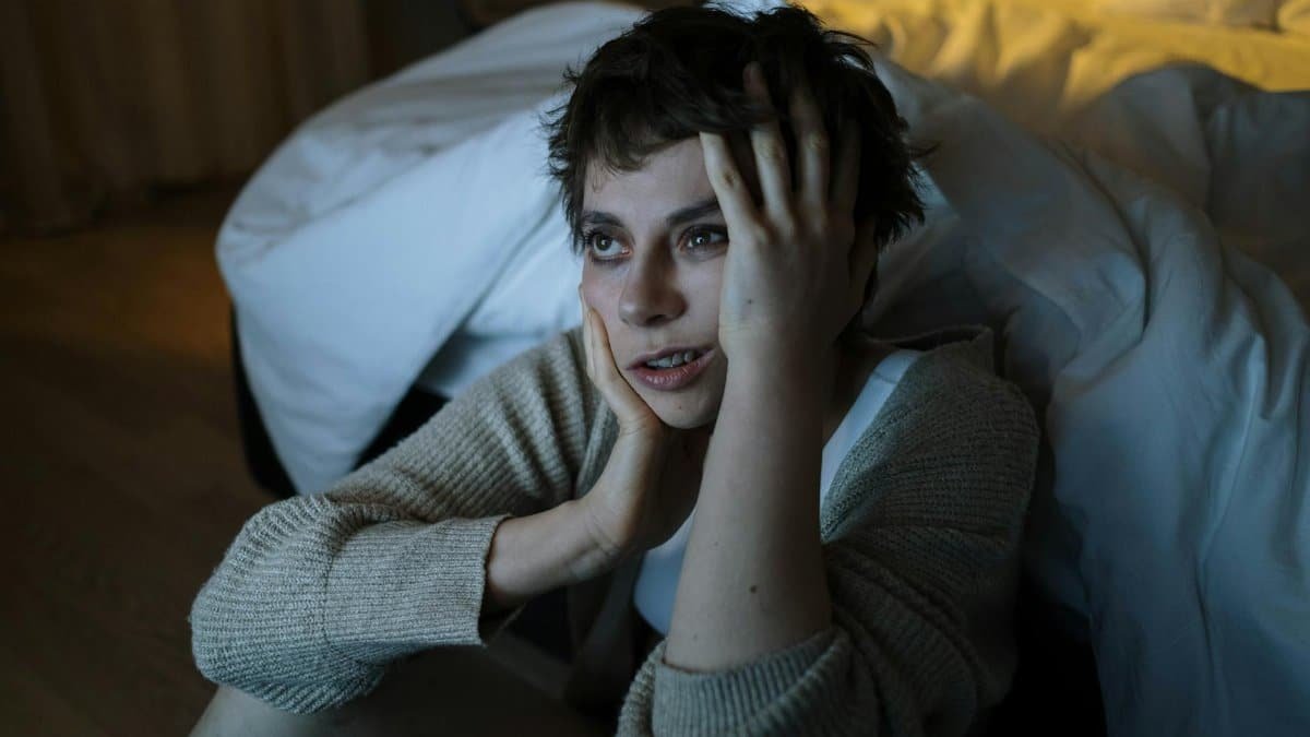 A woman sitting on the floor by a bed with a worried expression, touching her face.