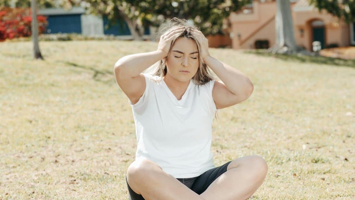 Young woman experiencing a headache while sitting on grass in a sunny park setting.