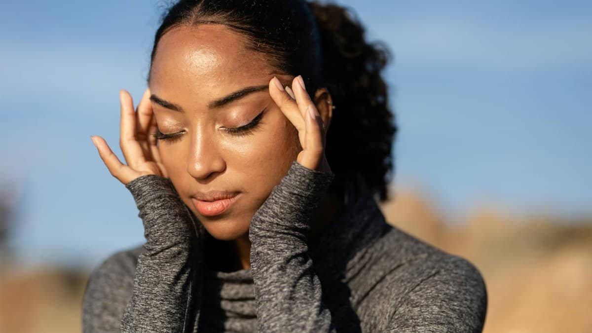 Close-up of a woman in a gray sweater holding her temples, experiencing a headache outdoors in the sunlight.