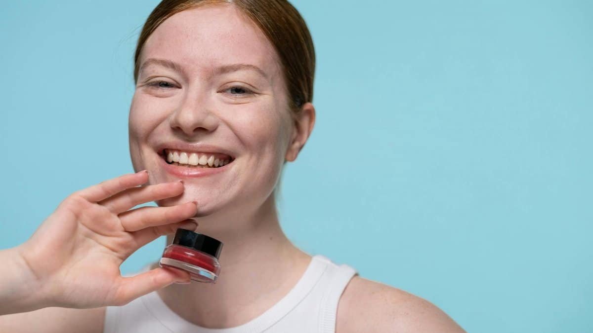 Cheerful woman holding a small tub of red cream against a blue background in a studio shoot.