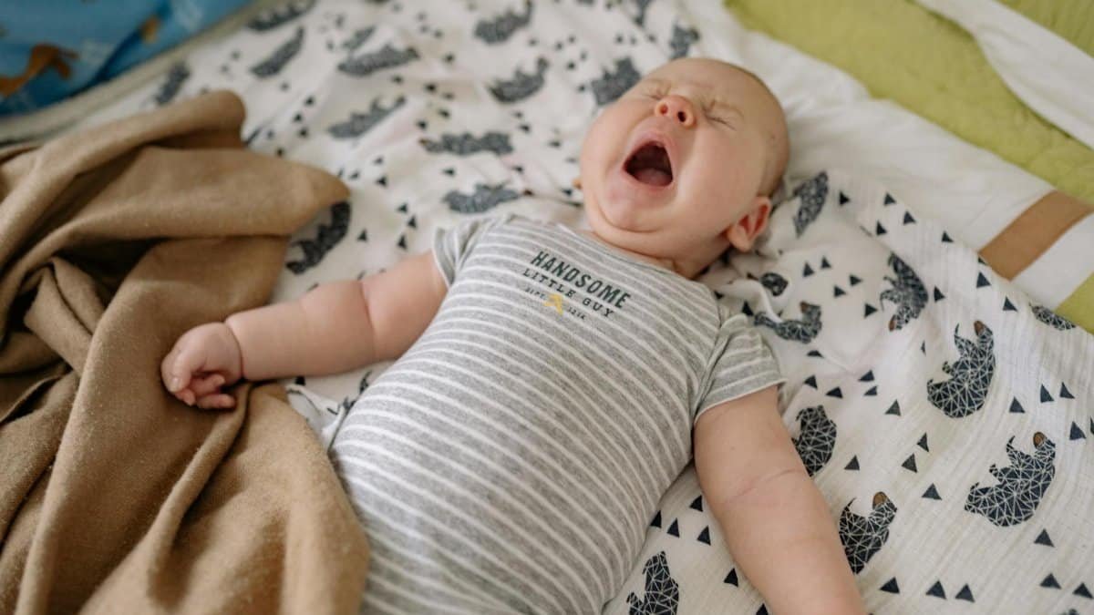 Cute baby in a striped onesie yawning on patterned bedding, cozy and relaxed.