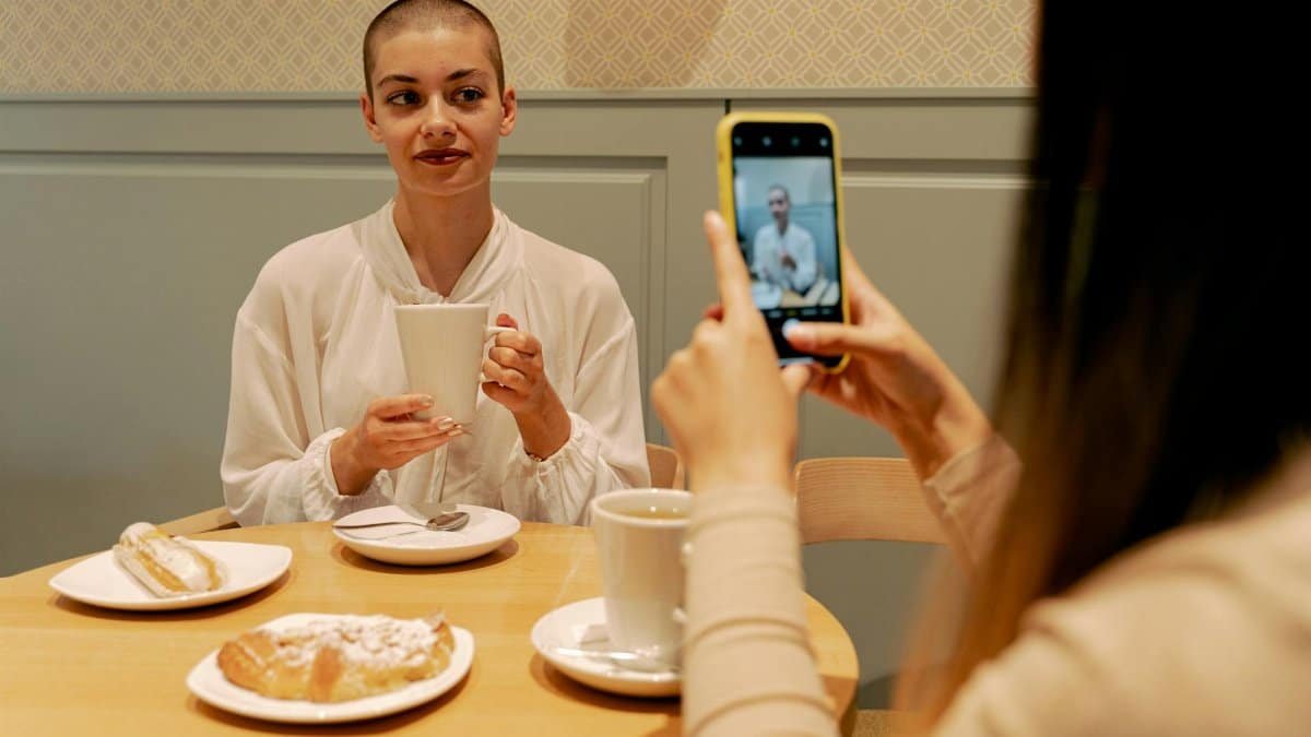 A woman with short hair enjoys coffee while a friend captures the moment. Indoor setting.