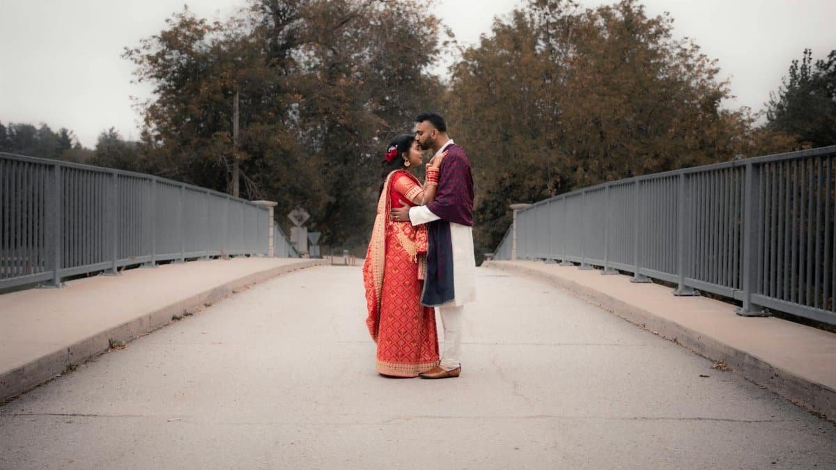 A couple in traditional clothing share a tender moment on a scenic bridge, surrounded by lush autumn trees.