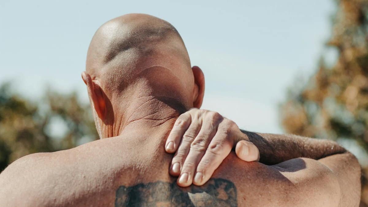 A bald man with a large tattoo massages his aching back outdoors, under clear skies.