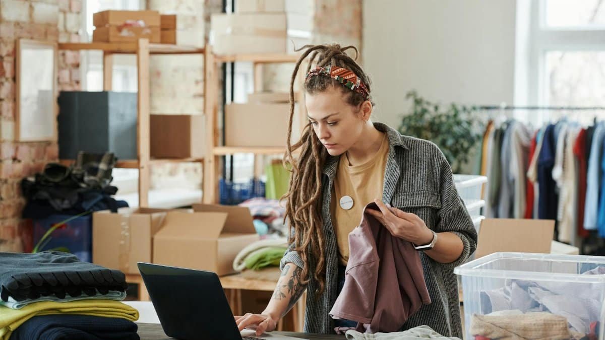 Woman with dreadlocks using a laptop while sorting clothes in a loft for volunteer work.