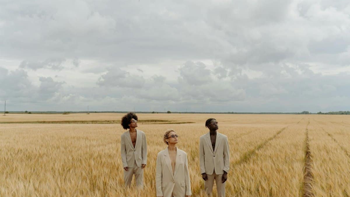 Three adults in stylish suits posing in a wide wheat field under a cloudy sky.