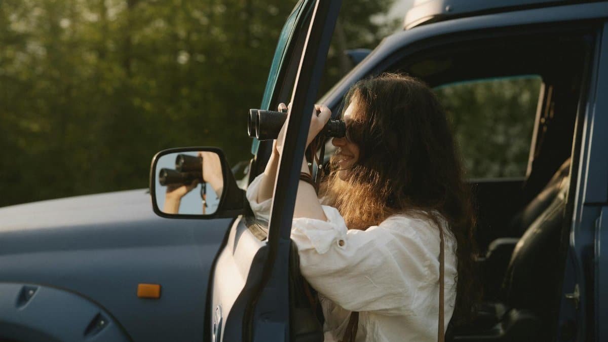 A woman using binoculars to observe nature from her car during a sunny day.