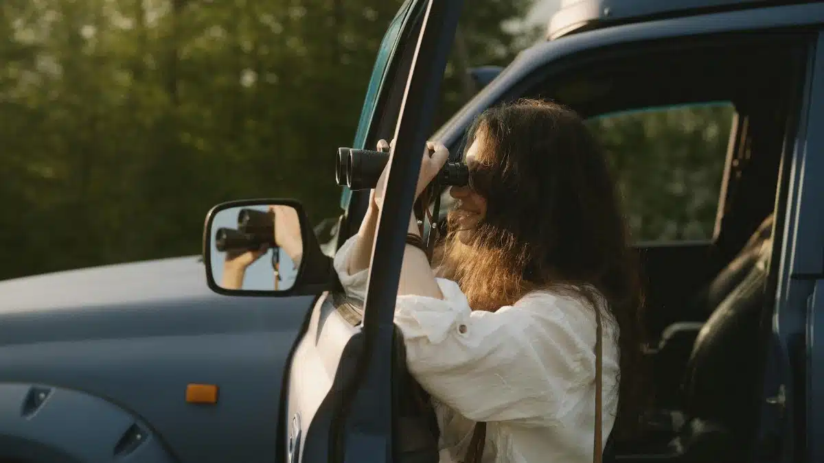 A woman using binoculars to observe nature from her car during a sunny day.