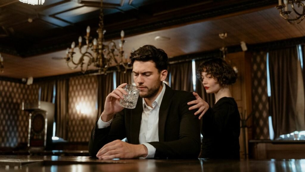An elegant bar scene with a thoughtful man holding a glass and a woman comforting him.