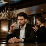 An elegant bar scene with a thoughtful man holding a glass and a woman comforting him.
