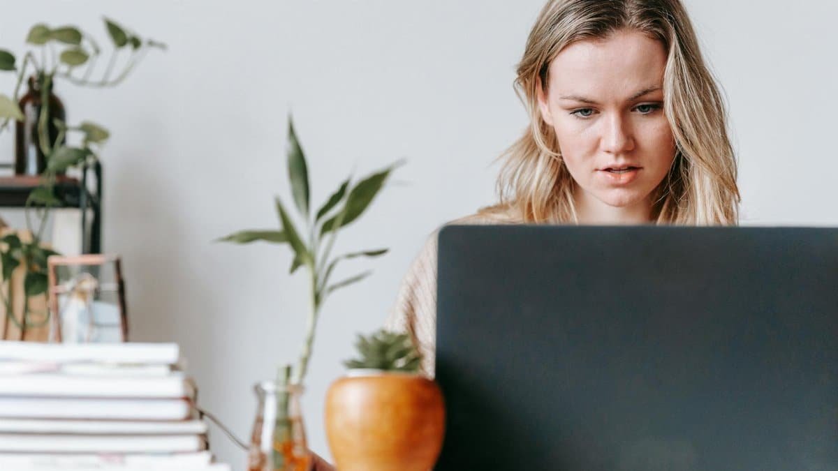 Young woman concentrating on her laptop in a home office setting, surrounded by houseplants and books.