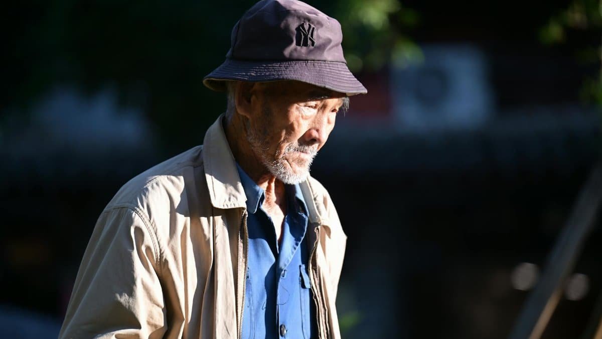 Elderly man in sunlit street wearing casual attire with a hat, deep in thought.
