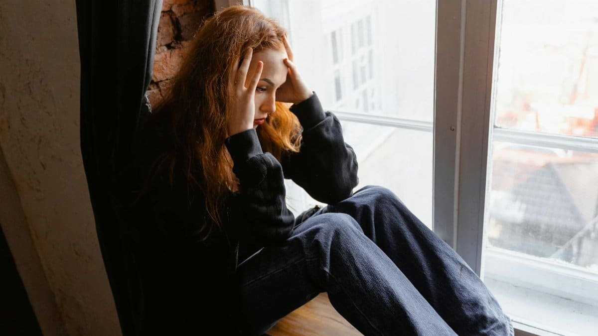 Woman sitting by a window, deep in thought, conveying emotions like distress and anxiety.