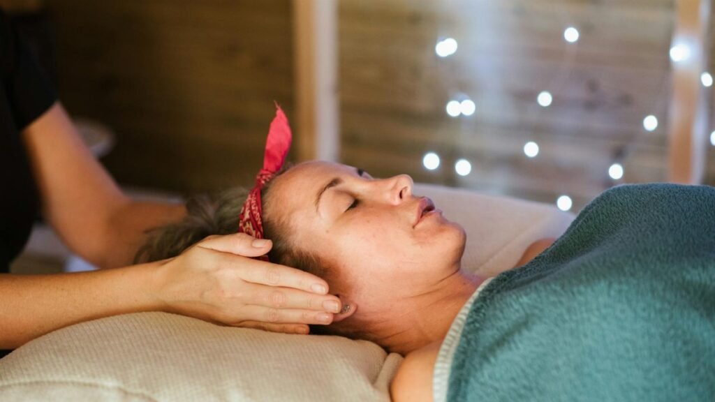 Side view of relaxed female patient with closed eyes lying on table under blanket while getting massage reiki during healing session in salon