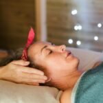 Side view of relaxed female patient with closed eyes lying on table under blanket while getting massage reiki during healing session in salon